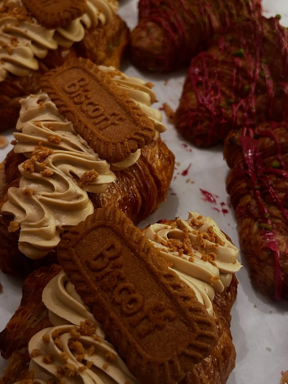 Kruasāni ar Biscoff krēmu un cepumu, aizmugurē kruasāni ar rozā glazūru un pistācijām