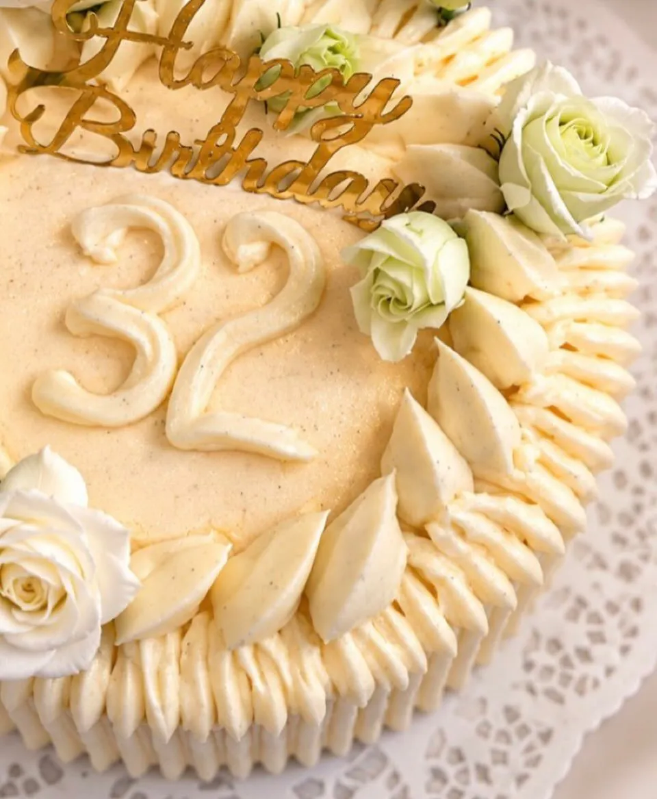 Svētku kūka ar krēma rotājumiem, baltām rozēm un zeltītu „Happy Birthday” uzrakstu