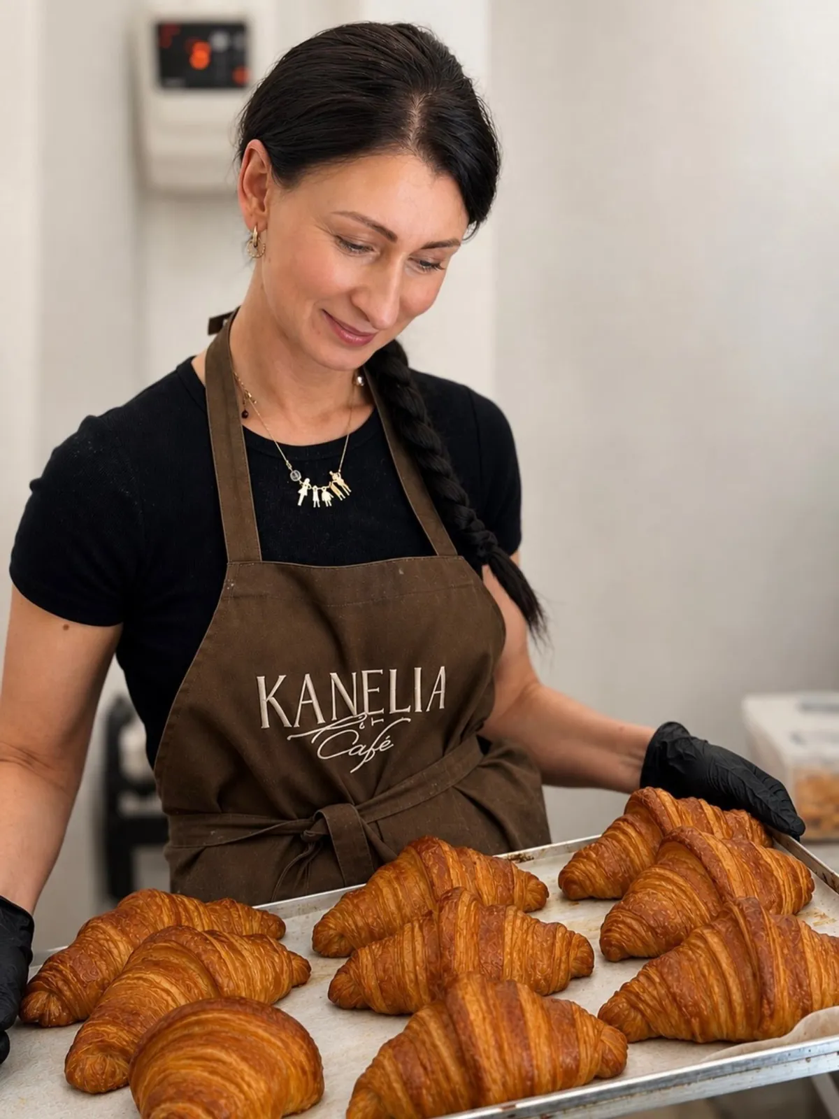 Konditore brūnā Kanelia Cafe priekšautā tur cepamo plāti ar svaigi ceptiem kruasāniem